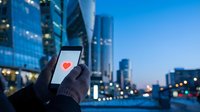 Person holding smartphone with red heart icon on screen against blurred city skyline at dusk, representing online dating and digital romance connections.