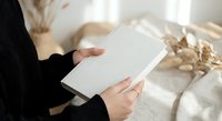 Woman in black sweater reading blank white book or magazine in cozy bedroom setting with soft natural lighting and neutral home decor elements.
