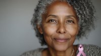 Close-up portrait of mature African American woman with natural gray curly hair wearing gray sweater and pink awareness ribbon against neutral background.