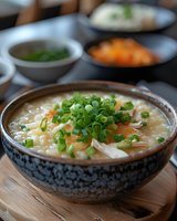 Asian rice porridge garnished with fresh green onions and julienned carrots in traditional ceramic bowl, served on bamboo mat with condiments in background.