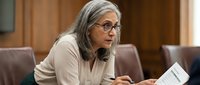 Senior woman with gray hair and glasses wearing beige blazer, holding document at conference table during business meeting or presentation.