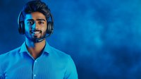 Young South Asian male customer service representative wearing headset and blue shirt smiling against dark blue background with dramatic lighting.