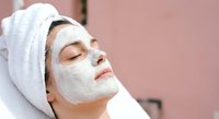 Young woman relaxing with white facial mask and towel wrapped around head during spa treatment for skincare routine and wellness therapy.