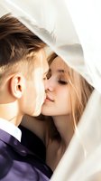 Young couple kissing under white veil, groom in purple suit and bride with light brown hair, romantic wedding moment with soft natural lighting.