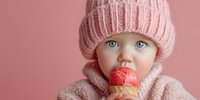 Adorable baby with blue eyes wearing pink knit hat and sweater eating strawberry ice cream cone against soft pink background for lifestyle content.