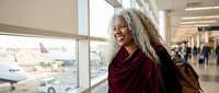 Smiling mature African American woman with long gray hair at airport terminal window overlooking planes on tarmac, ready for travel adventure.