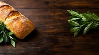 Fresh baked crusty artisan bread loaf with golden brown crust and green herbs on dark rustic wooden background with copy space for menu or recipe.