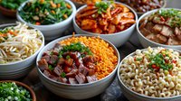 Korean food bowls with various traditional side dishes including kimchi, bean sprouts, marinated meat, noodles and vegetables served in ceramic bowls.