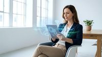 Woman in business casual attire holding tablet with digital holographic interface displaying data analytics and technology icons in bright modern office space with natural light.