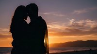Silhouette of couple kissing at sunset over ocean with golden sky and mountains, romantic moment for love and relationship content.