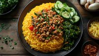 Savory minced meat with vegetables served over bright yellow rice, accompanied by fresh cucumber slices and sauteed greens on dark plate, rustic wooden surface.