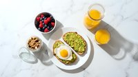 Healthy breakfast spread with avocado toast topped with fried egg, fresh berries, granola and orange juice on white marble surface for morning meal concepts.