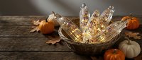Autumn harvest display with colorful corn ears in wicker basket surrounded by pumpkins and fall leaves on rustic wooden table for seasonal decoration.