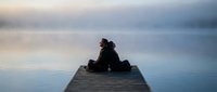 Silhouette of person sitting on wooden dock over calm lake water during peaceful sunset or sunrise with soft cloudy sky reflecting on surface for meditation concepts.