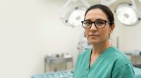 Female surgeon in green scrubs and glasses standing confidently in modern operating room with surgical lights and medical equipment in background.