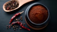 Ground coffee powder in black container with scattered peppercorns and dried red chili peppers on dark background, moody food photography composition.
