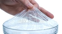 Close-up of hand stretching clear plastic wrap over glass bowl with water droplets, isolated on white background for food storage and kitchen preparation concepts.