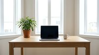 Modern minimalist home office workspace with laptop computer, coffee mug and green plant on wooden desk near bright windows for remote work productivity.