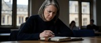 Middle-aged Caucasian woman with gray hair reading book in library, concentrated expression, natural lighting from windows, academic study environment.