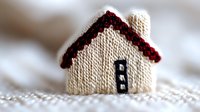 Miniature knitted house ornament with red roof and black door on white textured background, cozy home decor element for winter holiday season.
