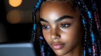Young African American woman with colorful braids working on laptop in modern office environment with warm bokeh lighting effects.