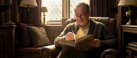 Elderly Caucasian man with glasses reading book in cozy armchair by window. Senior gentleman enjoying literature in comfortable home library setting.