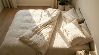 Unmade bed with crumpled beige linen sheets and pillows in morning sunlight, overhead view of bedroom interior with wooden floor and potted plant.