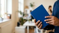 Man holding blue notebook in modern office workspace with colleagues working in background and natural light from windows.