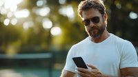 Confident mature Caucasian bearded man in white t-shirt and sunglasses using smartphone outdoors during golden hour sunset with natural bokeh background.
