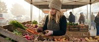 Young blonde woman in knit beanie shopping for fresh organic vegetables at outdoor farmers market stall with wooden crates and seasonal produce display.