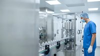 Pharmaceutical technician in blue scrubs and surgical cap operating automated laboratory equipment in sterile manufacturing facility for medical production and quality control.
