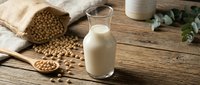 Fresh soy milk in glass with soybeans spilling from burlap sack on rustic wooden table. Healthy plant-based dairy alternative for vegan lifestyle and nutrition.