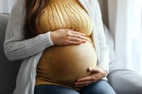 Pregnant woman in yellow sweater gently holding her baby bump, wearing cozy gray cardigan. Tender moment of motherhood and anticipation captured in soft natural light.