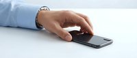Hand in blue shirt sleeve reaching for smartphone lying on white surface, close-up view for business and technology concepts.