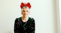 Blonde woman wearing festive red bow headband and dark sequined sweater, smiling at camera against white background for holiday portraits and seasonal marketing campaigns.