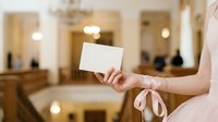 Woman in light pink dress holding blank white card with ribbon in luxury hotel lobby or upscale interior space for greeting, invitation, or promotional messaging.