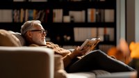 Senior Caucasian man with gray beard and glasses relaxing on couch reading book in cozy home library with warm lighting and bookshelves in background.