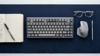 Modern minimalist workspace with wireless keyboard, mouse, notebook and glasses on dark blue desk surface for productivity and office design.