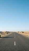 Straight asphalt road with white center line stretching through arid desert landscape under clear blue sky, depicting travel, journey, and remote highway exploration.