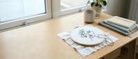 Hand embroidery hoop with floral design on wooden desk near window with coffee cup and books creating cozy crafting workspace for needlework projects.