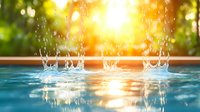 Splashing water drops creating crown shapes in swimming pool at sunset, sun rays reflecting on blue rippled surface with bokeh effect background.