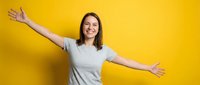 Young woman with long dark hair and fair complexion, wearing a light gray t-shirt, smiling with arms outstretched in a welcoming gesture against a bright yellow background. Suitable for lifestyle, happiness, and positivity concepts.