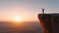 Silhouette of person with raised arms celebrating success on mountain cliff edge during golden sunrise over desert landscape for motivation concepts.