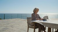 Senior woman working on laptop at outdoor terrace table overlooking ocean, remote work lifestyle concept for business and retirement planning.