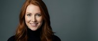 Young redhead businesswoman smiling confidently in black turtleneck against gray background for corporate headshot and professional portrait photography.