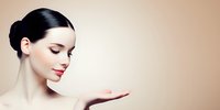 Elegant woman with sleek hair bun and red lips presenting product on open palm against neutral beige background, perfect for beauty and skincare advertising.