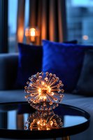 Modern spherical crystal light fixture reflecting on glass table with blue velvet pillows in background creating cozy evening atmosphere.