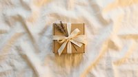 Wrapped gift box with golden ribbon bow on neutral fabric background with natural shadows and sunlight.