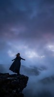 Silhouette of woman standing on rocky cliff edge with arms outstretched against dramatic twilight sky with purple and blue clouds for freedom and adventure concepts.