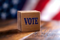 Wooden block with vote text against blurred American flag background on rustic wooden surface, representing democracy and electoral process.